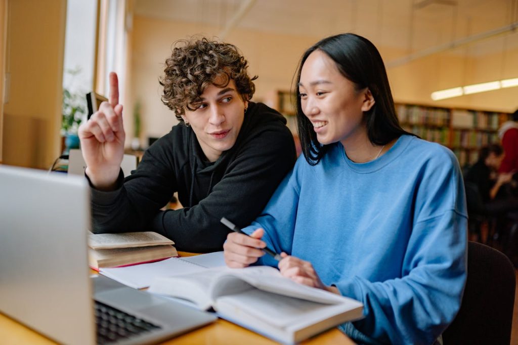 Two students collaborating on a study project in the library, using a laptop and books.