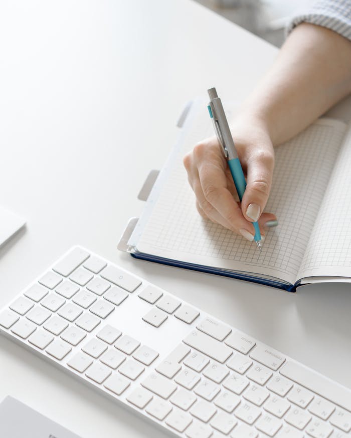 Close-up of a hand writing in a notebook next to a white keyboard on a desk.